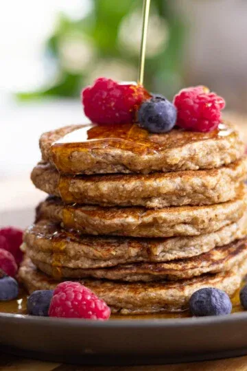 Stack of banana oatmeal pancakes with syrup, raspberries, and blueberries on a plate.
