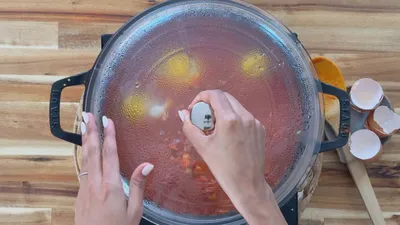 Person lifting lid from pot cooking eggs and vegetables on stove, wooden spoon and eggshells beside.