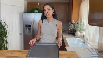 Woman in a kitchen holding a baking tray, standing by a wooden counter and stainless steel fridge.
