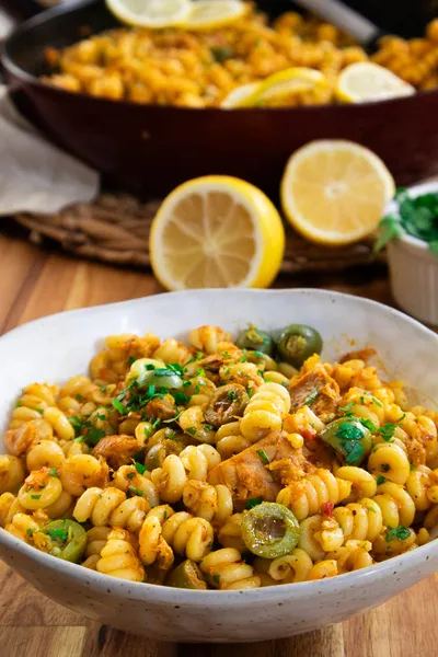 Bowl of spiral pasta with olives and herbs, garnished with lemon slices on a wooden table.