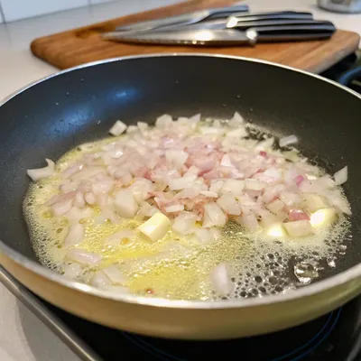 Chopped shallots frying in a pan with butter on a stove, cutting board and knives in the background.