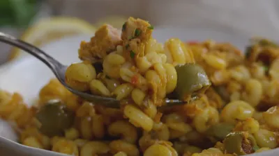 Close-up of fork with hearty pasta, featuring olives, tuna, and herbs.