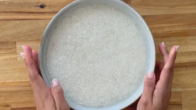 Hands holding a bowl of rice soaking in water on a wooden countertop.