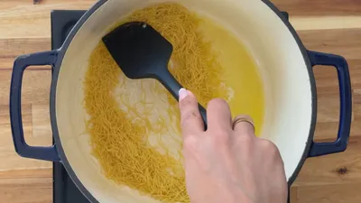 Hand stirring thin pasta in a pot with a black spatula on a wooden surface.