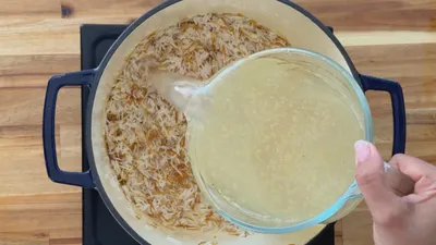 Pouring broth into a pot of rice on a wooden surface.