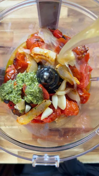 Food processor with tomatoes, pesto, garlic, and onions being blended from above.