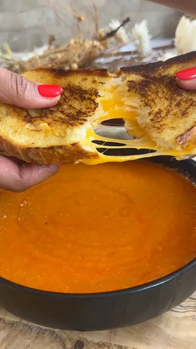 Grilled cheese being pulled apart above a bowl of tomato soup.