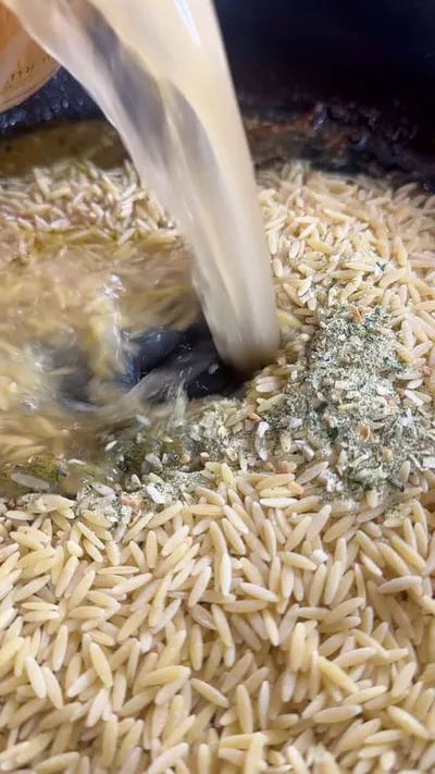 Pouring broth into orzo pasta with seasoning in a pan.