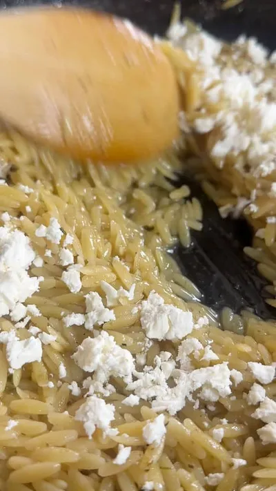 Orzo pasta with crumbled feta cheese being stirred with a wooden spoon in a pan.