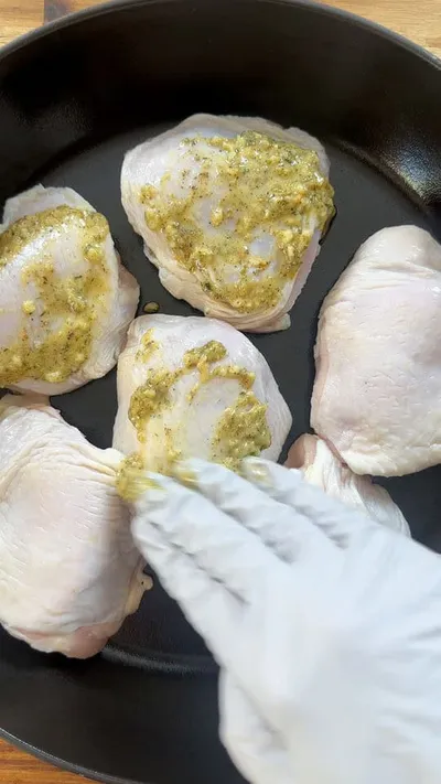 Gloved hand applying seasoning on raw chicken thighs in a pan.