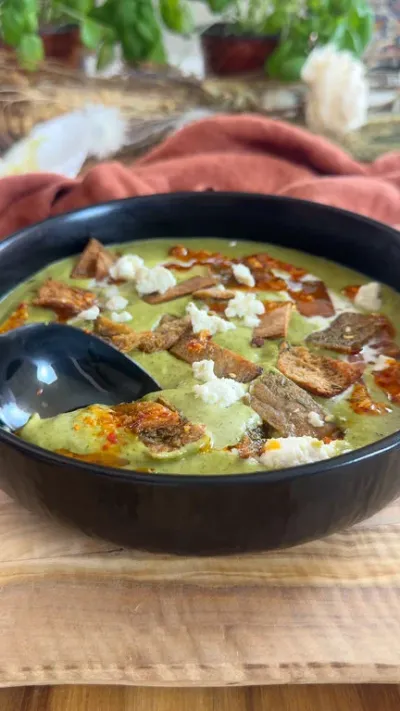 Creamy green soup topped with pita chips and cheese in a black bowl on a wooden board.