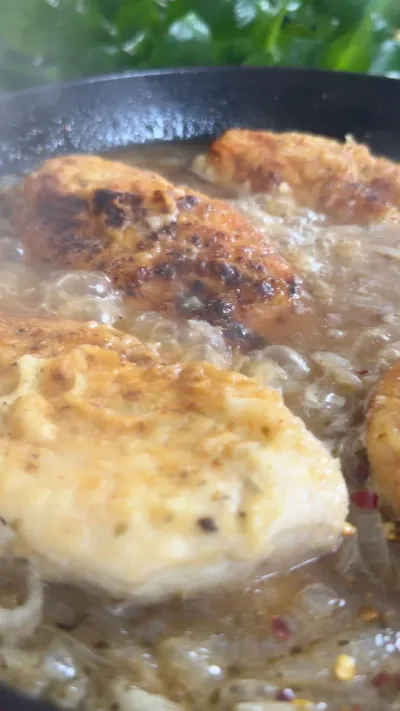 Chicken breasts sizzling in a pan with onions and spices, surrounded by fresh herbs.