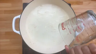 Pouring liquid from a measuring cup into a white pot on a stovetop.