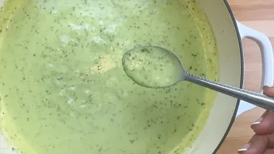 Spoon dipping into a pot of creamy pesto sauce on a wooden countertop.