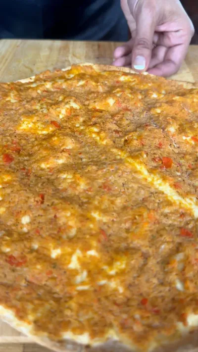 Close-up of a hand holding lahmacun with a minced meat topping on a wooden board.