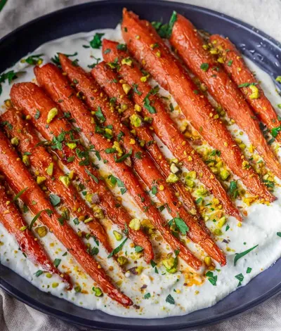 Roasted Harissa Maple Carrots on whipped feta, garnished with pistachios and herbs, served on a dark oval plate.