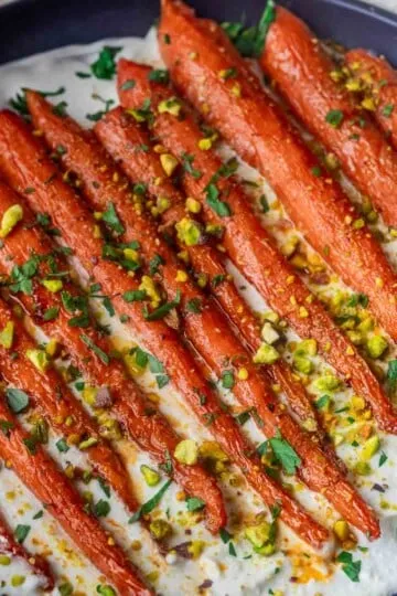 Roasted Harissa Maple Carrots, chopped pistachios and herbs on creamy sauce in a dark dish.