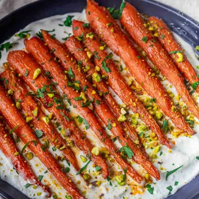 Roasted Harissa Maple Carrots, chopped pistachios and herbs on creamy sauce in a dark dish.