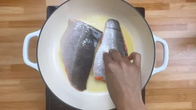 Hand placing salmon fillets in a white pan on a wooden surface.