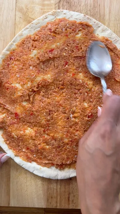 Spreading meat on flatbread with a spoon on a wooden surface.