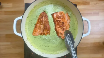 Two salmon fillets in creamy pesto sauce in a white pan on wooden counter, with tongs holding one fillet.