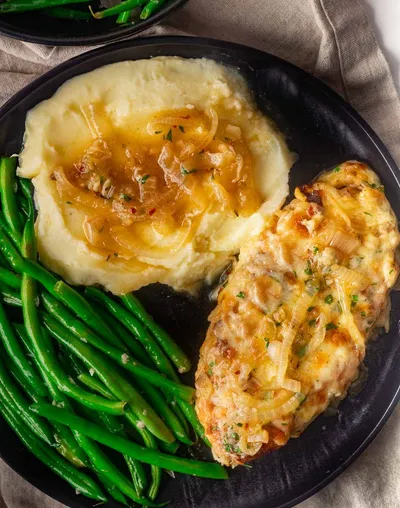French onion soup chicken with caramelized onions, mashed potatoes, and green beans on a black plate.
