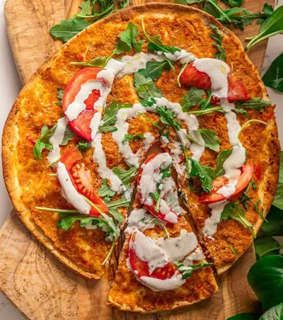 Thin crust pizza topped with arugula, tomato slices, and ranch dressing on a wooden board.