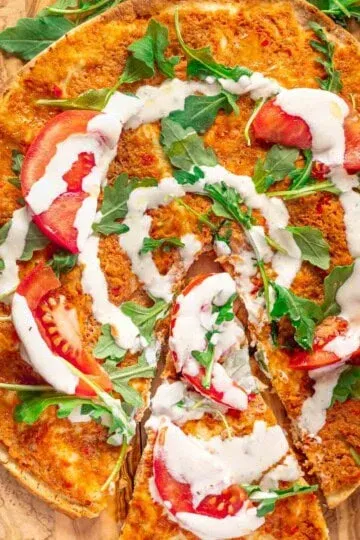 Lebanese chicken pizza with tomato slices, arugula, and white sauce drizzle on a wooden board.
