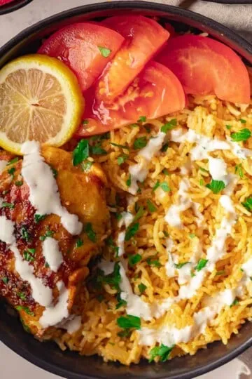 Grilled chicken with yogurt sauce, yellow rice, tomato slices, and a lemon wedge in a black bowl.