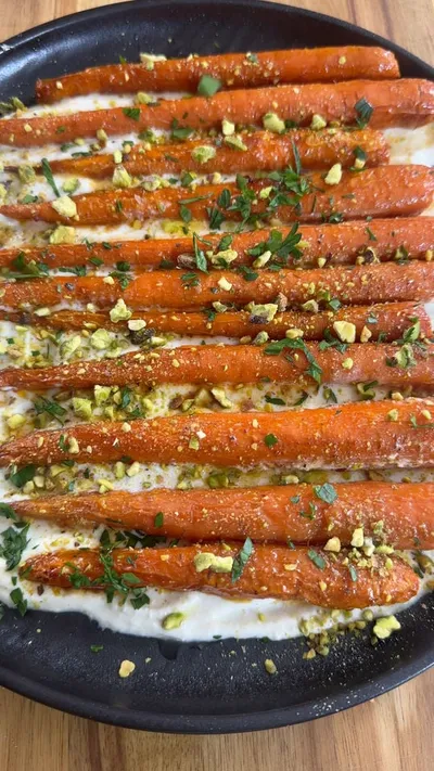 Roasted carrots with crushed pistachios and herbs on a bed of creamy sauce in a black dish.