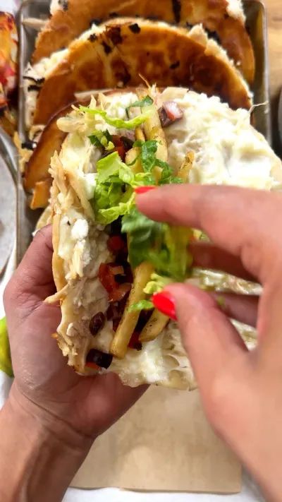 Person holding a taco with lettuce, chicken, cheese, and vegetables, with tacos in the background.