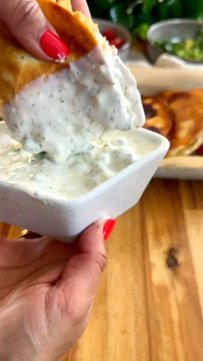 Hand holding a taco dipping into white sauce in a small bowl on a wooden table.