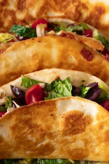 Crispy tacos with lettuce, tomatoes, olives, and melted cheese in a folded tortilla shell.