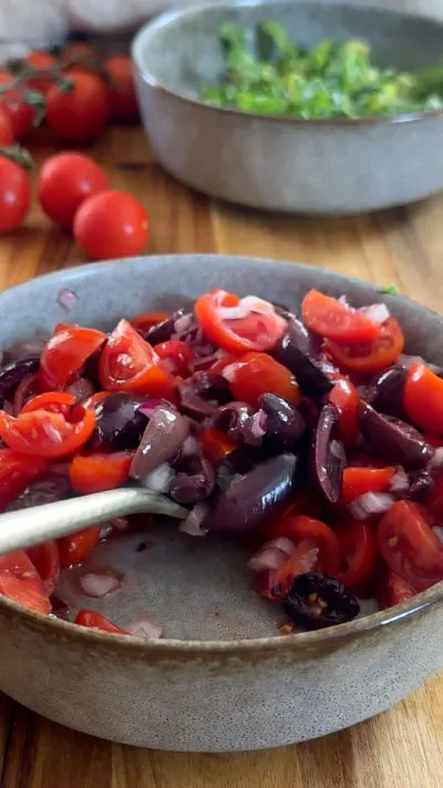 Chopped tomatoes and olives with onions in a bowl, with cherry tomatoes and greens in the background.