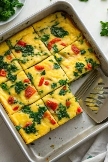 Sheet pan eggs with spinach and tomatoes in a square pan, partially sliced, metal spatula on the side.
