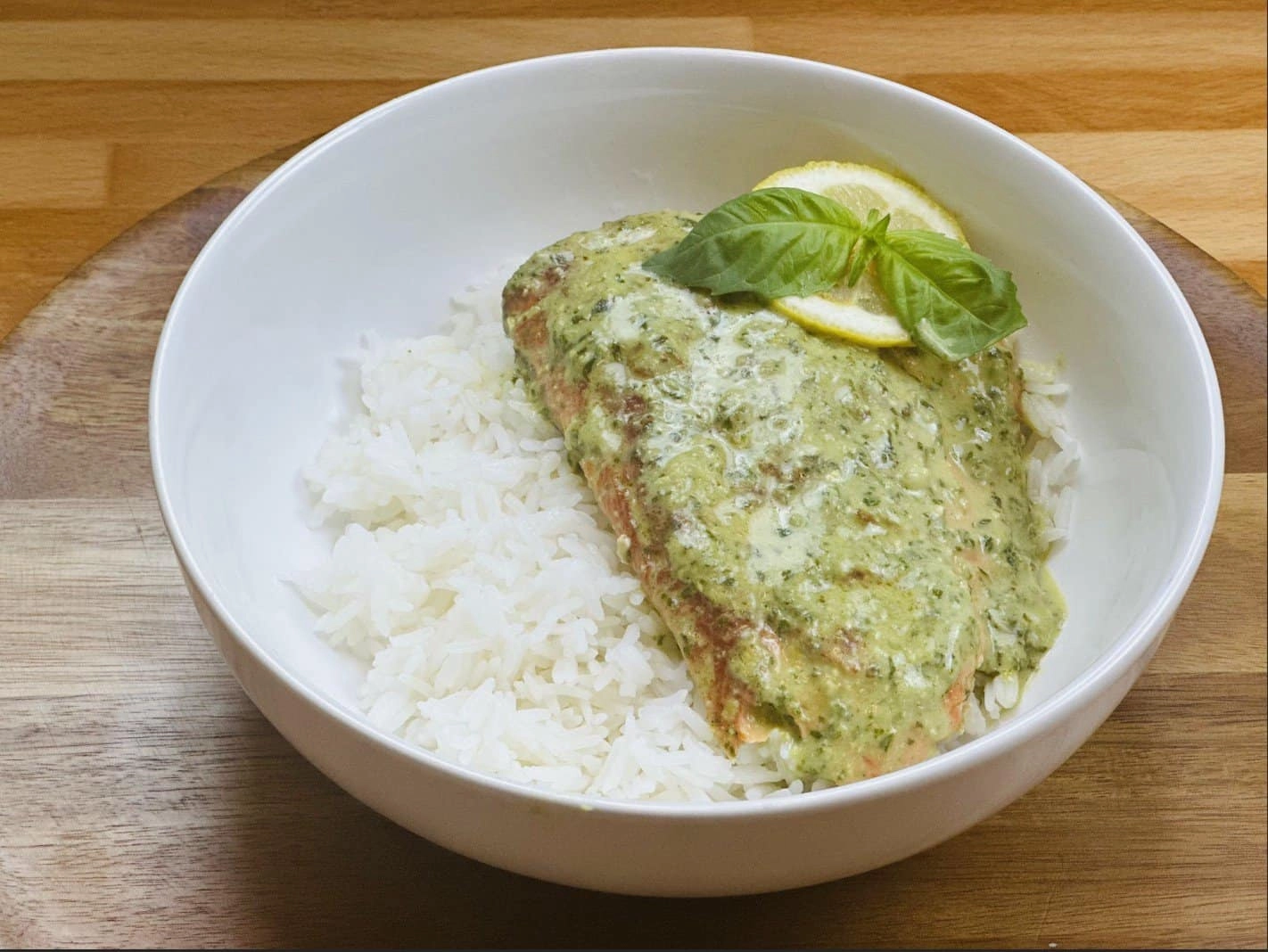 Salmon with pesto sauce on rice, garnished with basil and lemon, served in a white bowl on a wooden table.