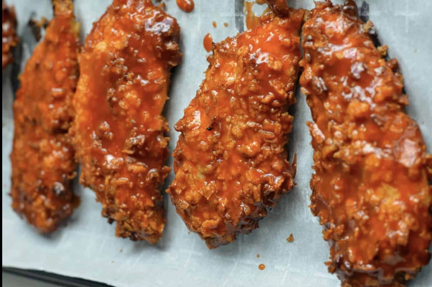 Crispy fried chicken tenders coated in spicy sauce on parchment paper.