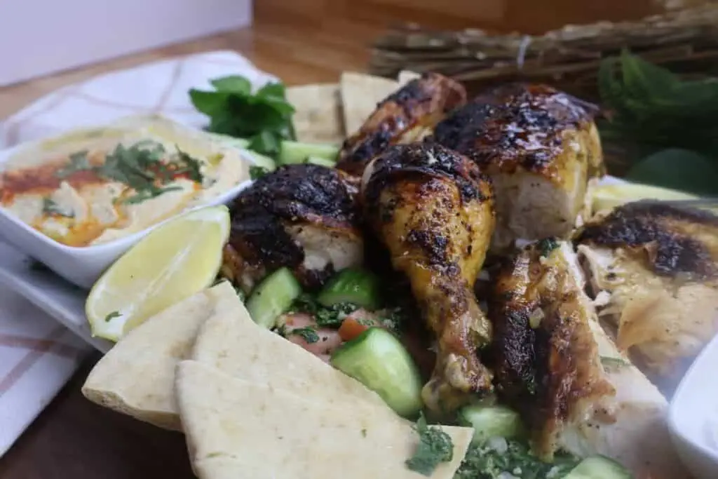 Grilled chicken platter with hummus, pita bread, cucumber salad, and lemon wedges on a white plate.
