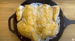 Whole chicken marinated with spices on a bed of sliced onions in a cast iron skillet, ready for roasting.