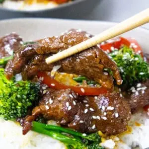 Beef stir-fry with broccoli, red peppers, and sesame seeds on rice, held with chopsticks. Asian cuisine dish.