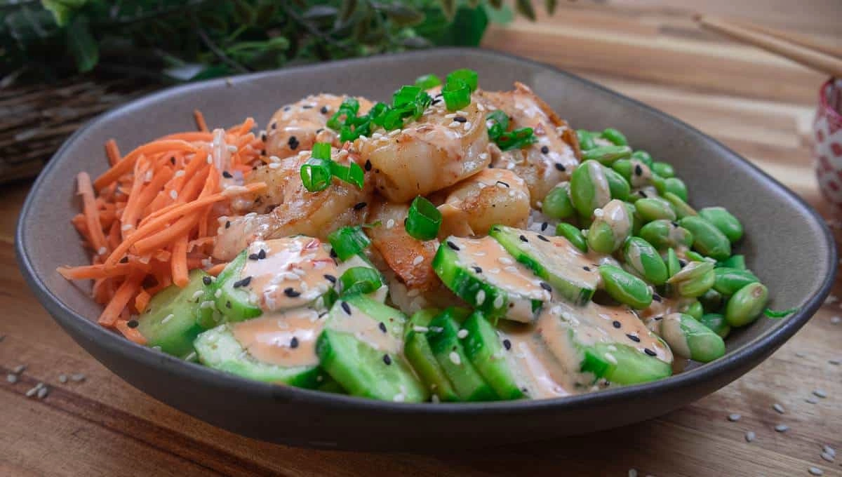 Colorful shrimp poke bowl with carrots, edamame, cucumbers, and spicy mayo dressing on a wooden table.