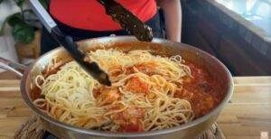 Delicious spaghetti with rich tomato sauce in a skillet being stirred with tongs on a wooden counter.