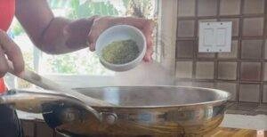 Person adding herbs to a steaming pan in a kitchen setting.