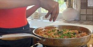 Person cooking spaghetti in a pan with vegetables, herbs in a cozy kitchen setting.