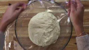 Person covers dough in a glass bowl with plastic wrap, preparing it to rise.