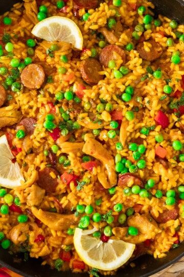Pan of vibrant paella with saffron rice, peas, chorizo, chicken, red peppers, and lemon wedges.