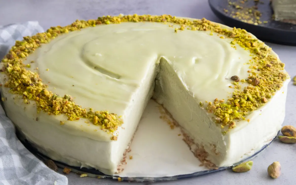 Creamy pistachio cake with a slice cut out, topped with crushed pistachios on a light gray surface.