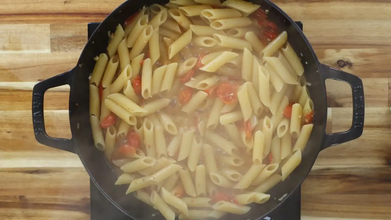 Steaming penne pasta with cherry tomatoes cooking in a black pot on a wooden surface.