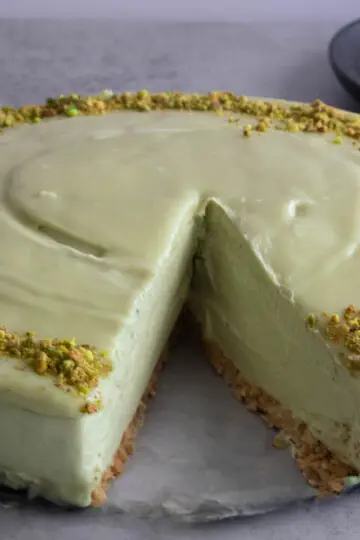 Creamy no bake pistachio cheesecake with a slice missing, topped with crushed pistachios on a gray surface.