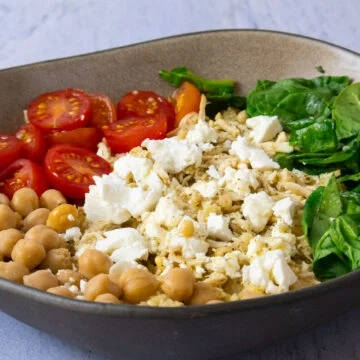 Bowl with chickpeas, cherry tomatoes, feta, spinach, and grains on a stone surface.